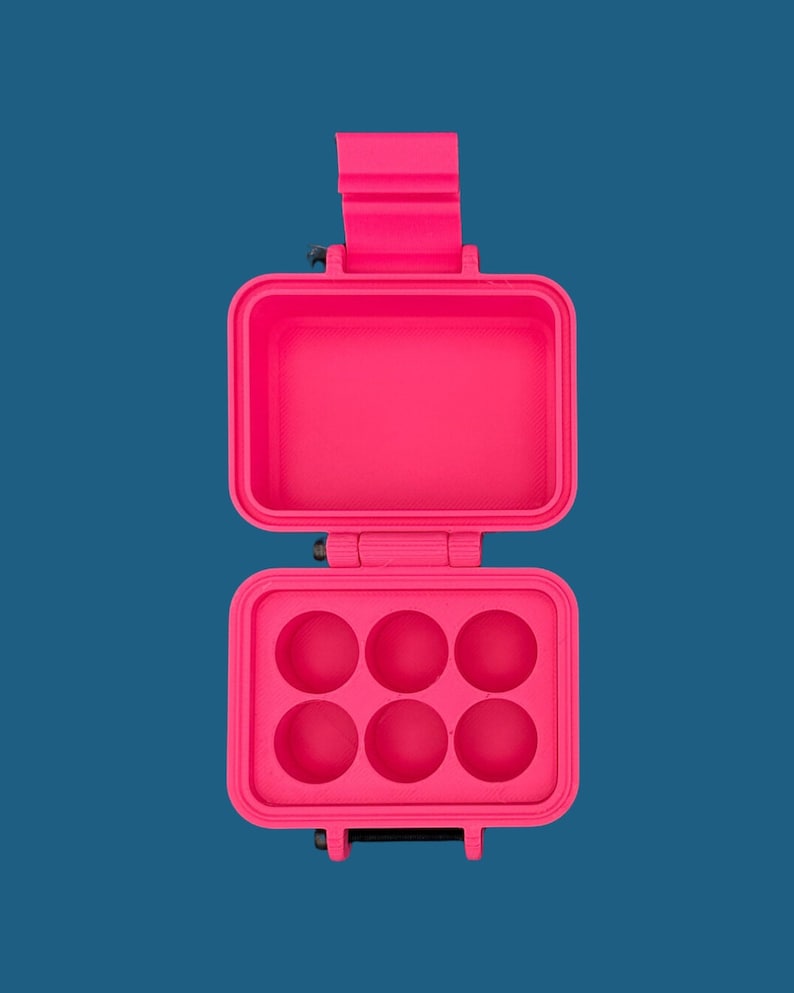 May include: A bright neon pink rectangular storage case with a hinged lid. The interior of the bottom section features six circular indentations arranged in two rows of three, while the top section is an open, empty rectangular compartment.