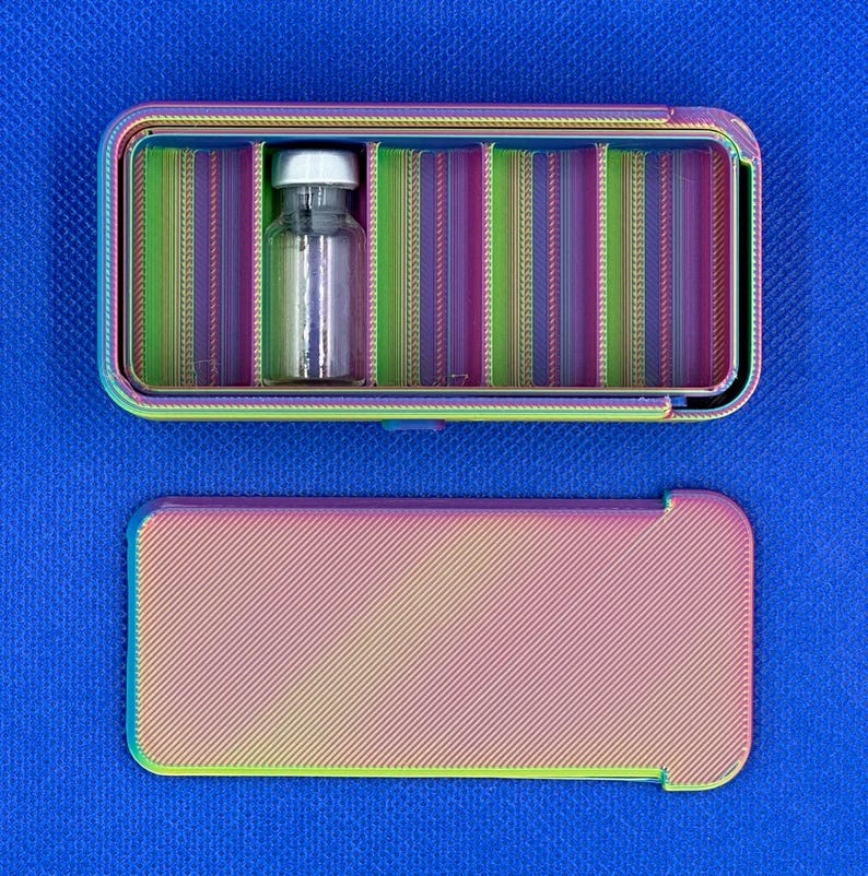 May include: A rectangular, iridescent, multi-colored storage case with a sliding lid. The interior features vertical dividers creating multiple compartments, with one clear glass vial containing a silver cap placed inside one section.
