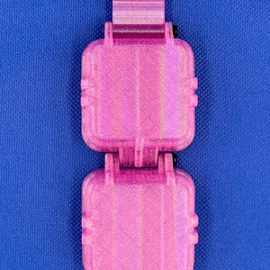 May include: A bright pink, 3D-printed, dual-compartment storage case with a textured, glittery finish. The rectangular unit features two hinged, square-shaped containers connected by a central hinge mechanism and secured with black metal screws.