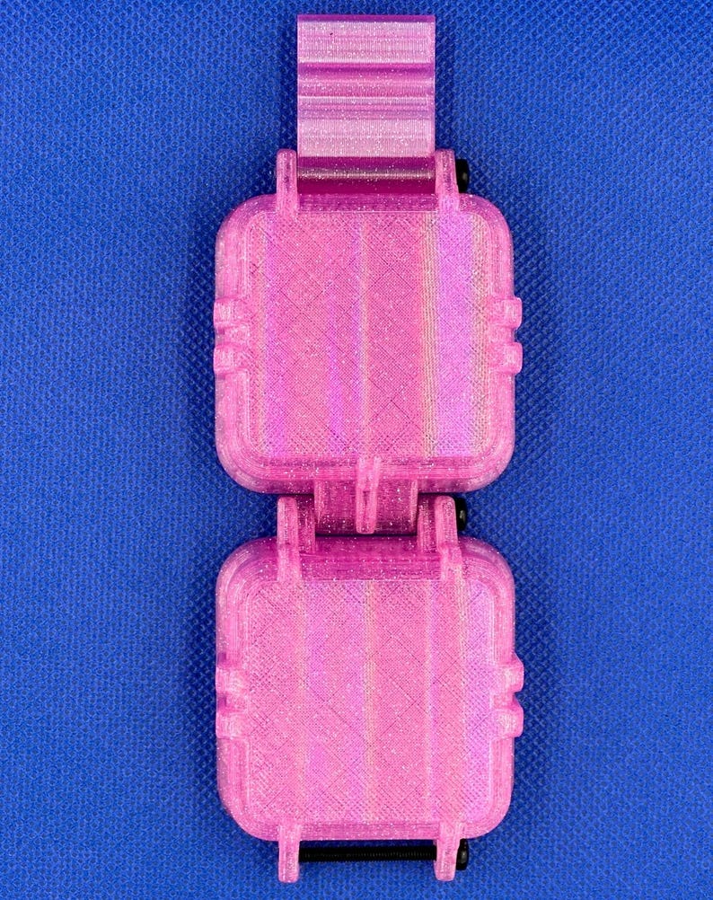 May include: A bright pink, 3D-printed, dual-compartment storage case with a textured, glittery finish. The rectangular unit features two hinged, square-shaped containers connected by a central hinge mechanism and secured with black metal screws.