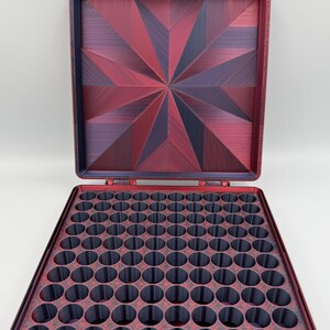 May include: A square storage case with a hinged lid, featuring a deep red and dark purple starburst pattern on the interior lid. The base contains a grid of numerous circular compartments in a dark gray finish, designed for organized storage.