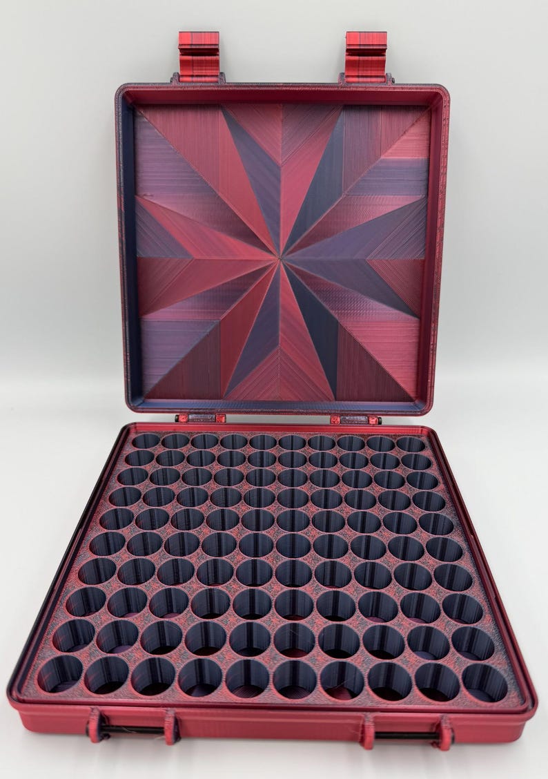 May include: A square storage case with a hinged lid, featuring a deep red and dark purple starburst pattern on the interior lid. The base contains a grid of numerous circular compartments in a dark gray finish, designed for organized storage.