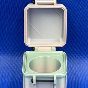 May include: A square, light-coloured, 3D-printed storage container with an open hinged lid. The base features a circular interior cutout and a black horizontal metal bolt fastener on the front. The exterior has a smooth, matte finish.