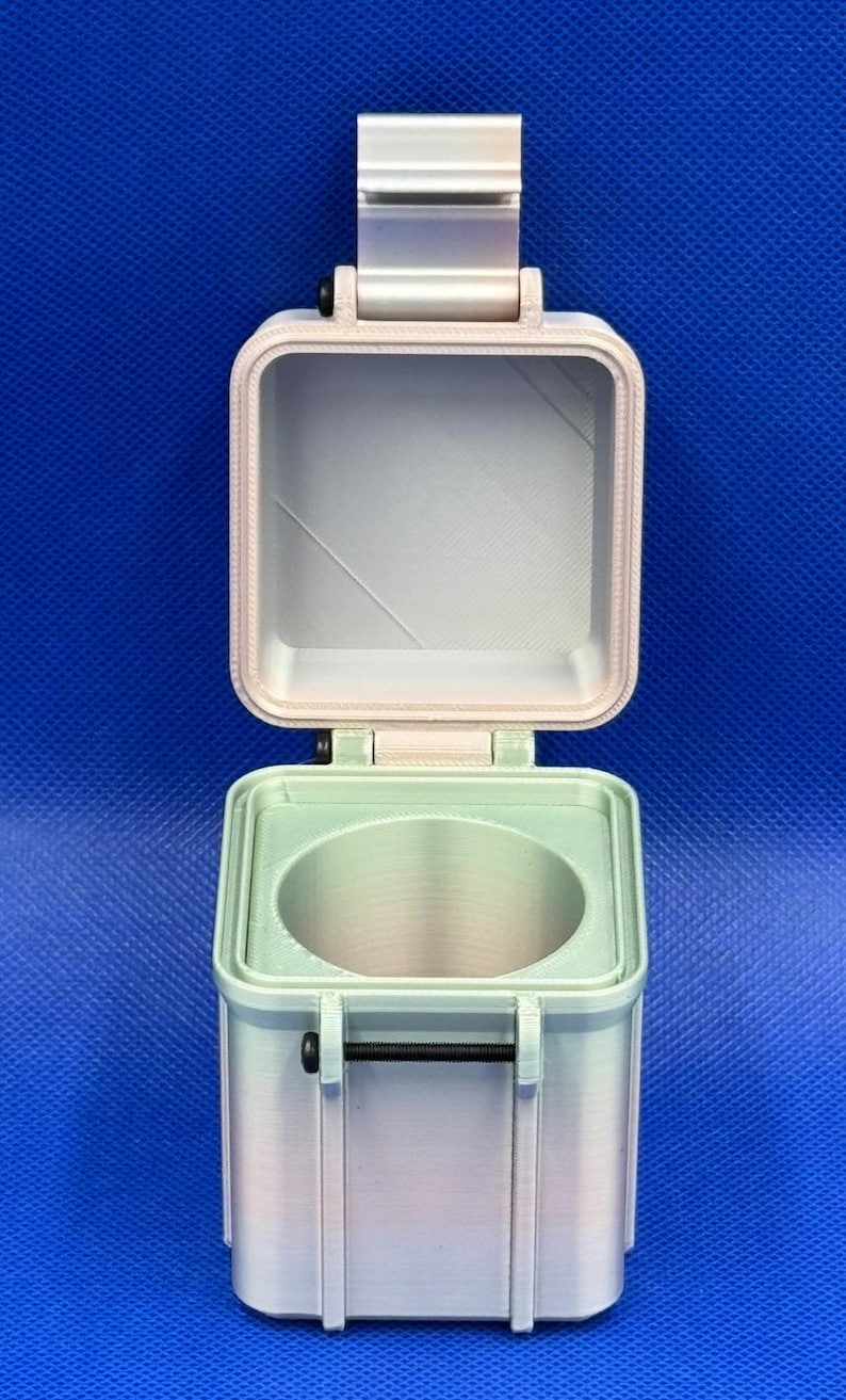 May include: A square, light-coloured, 3D-printed storage container with an open hinged lid. The base features a circular interior cutout and a black horizontal metal bolt fastener on the front. The exterior has a smooth, matte finish.
