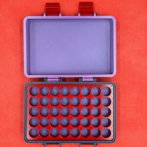 May include: Open, rectangular, two-tone storage box. The lid is a light purple, and the base is red with a grid of circular indentations. The box has two red clasps and is set against a red background.