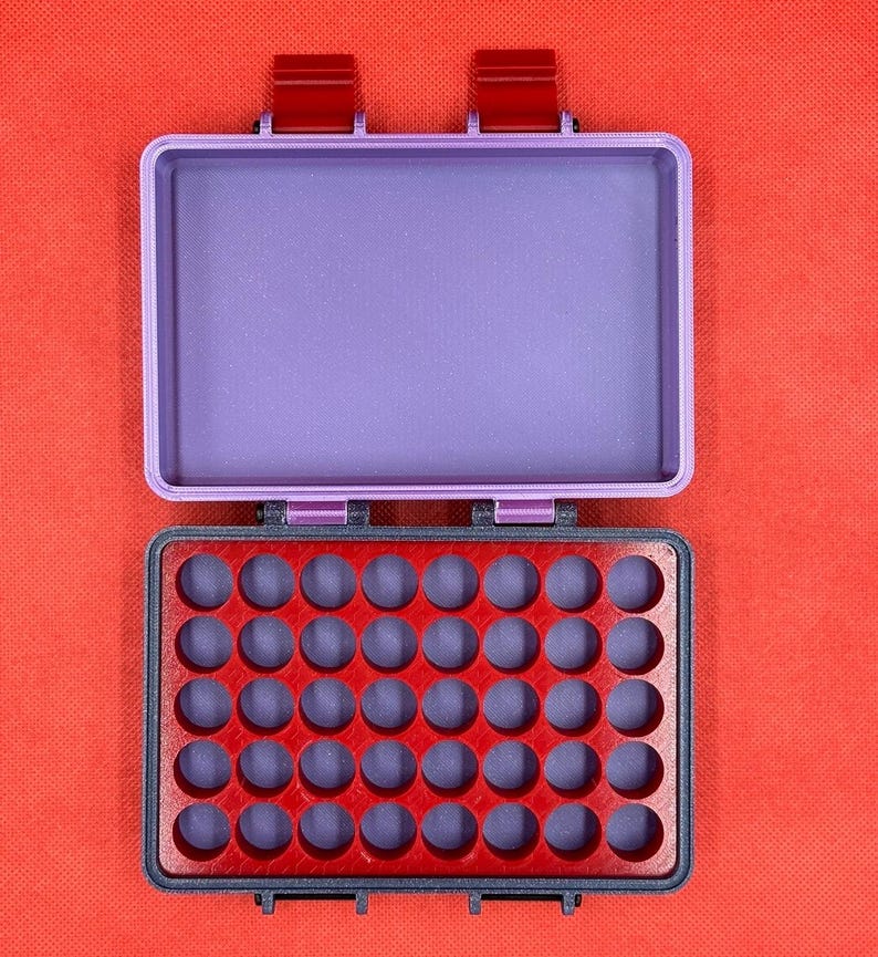 May include: Open, rectangular, two-tone storage box. The lid is a light purple, and the base is red with a grid of circular indentations. The box has two red clasps and is set against a red background.