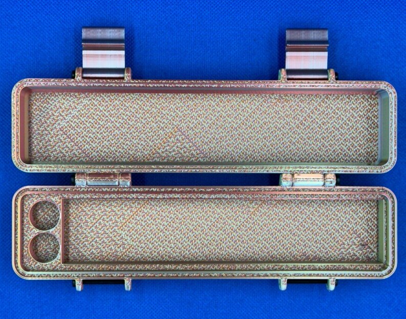 May include: A two-piece, brown and gold, rectangular storage case with a textured surface. The case has two compartments, one with two small circular holes and the other with a larger rectangular space.