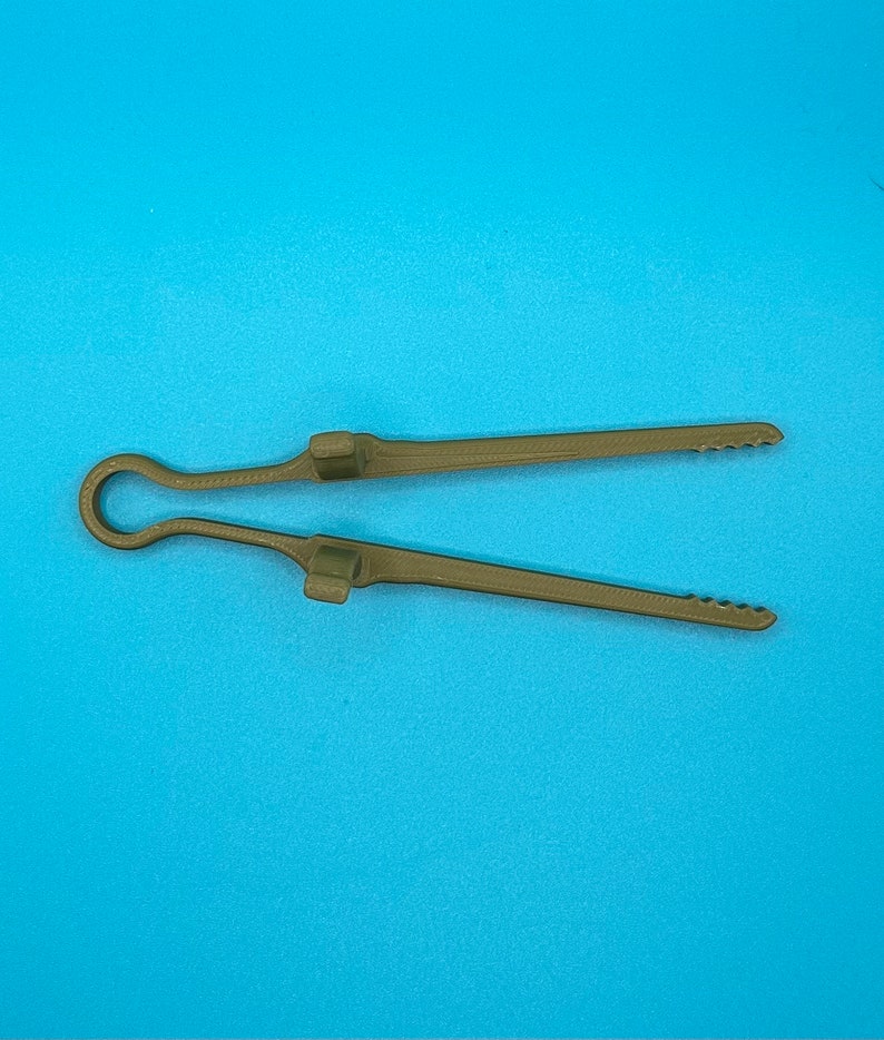 May include: A pair of olive green kitchen tongs featuring a single-piece construction with a looped handle, two raised rectangular grips along the center of each arm, and serrated edges at the tips for improved gripping of food items.