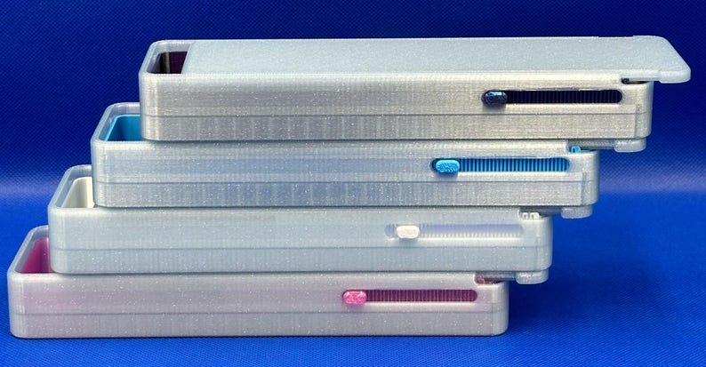May include: A stack of four rectangular, silver-toned plastic cases, each featuring a sliding mechanism on the side. The sliders are colored dark blue, light blue, white, and pink, matching the interior accents visible at the open ends.