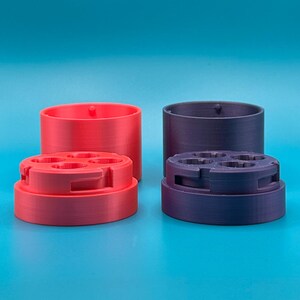 Op de afbeelding: Twee cilindrische houders, &eacute;&eacute;n rood en &eacute;&eacute;n donkerpaars, elk bestaande uit een basis met vier ronde openingen en een bijpassend deksel. De onderdelen zijn gemaakt van een mat materiaal met zichtbare 3D-geprinte lagen.