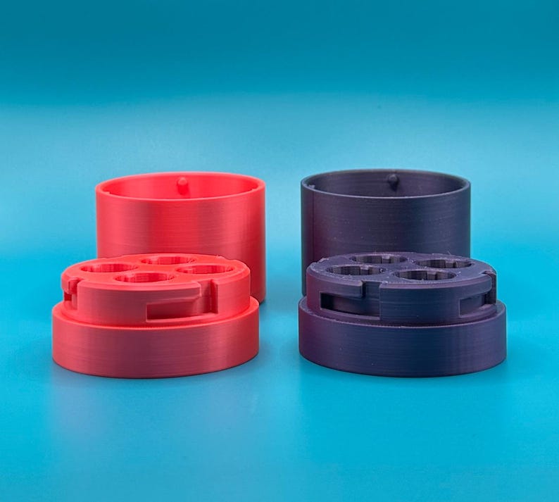 Op de afbeelding: Twee cilindrische houders, &eacute;&eacute;n rood en &eacute;&eacute;n donkerpaars, elk bestaande uit een basis met vier ronde openingen en een bijpassend deksel. De onderdelen zijn gemaakt van een mat materiaal met zichtbare 3D-geprinte lagen.