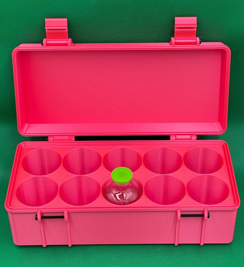 May include: A bright pink rectangular storage case with an open lid and ten circular compartments. One small clear glass vial with a vibrant lime green cap is placed inside one of the circular slots. The case features two black hinges on the front.