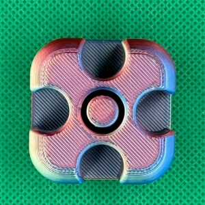 May include: A square-shaped fidget spinner with rounded corners, featuring a gradient color transition from blue to purple to red. The surface has a visible 3D-printed texture with diagonal lines and four circular cutouts surrounding a central cap.