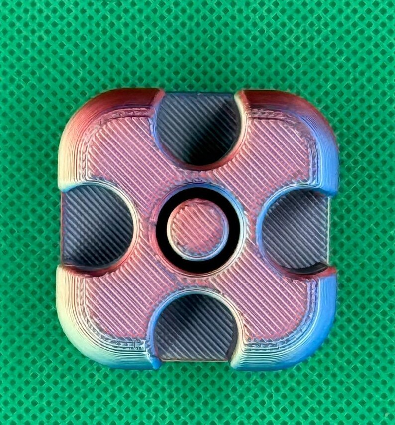 May include: A square-shaped fidget spinner with rounded corners, featuring a gradient color transition from blue to purple to red. The surface has a visible 3D-printed texture with diagonal lines and four circular cutouts surrounding a central cap.
