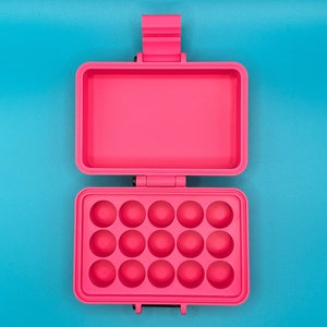 Puede incluir: Un estuche de almacenamiento rectangular de color rosa brillante con tapa abatible. El interior de la parte inferior presenta quince hendiduras circulares dispuestas en tres filas de cinco, mientras que la parte superior es hueca.