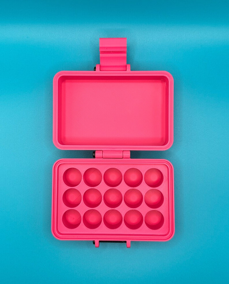 Puede incluir: Un estuche de almacenamiento rectangular de color rosa brillante con tapa abatible. El interior de la parte inferior presenta quince hendiduras circulares dispuestas en tres filas de cinco, mientras que la parte superior es hueca.