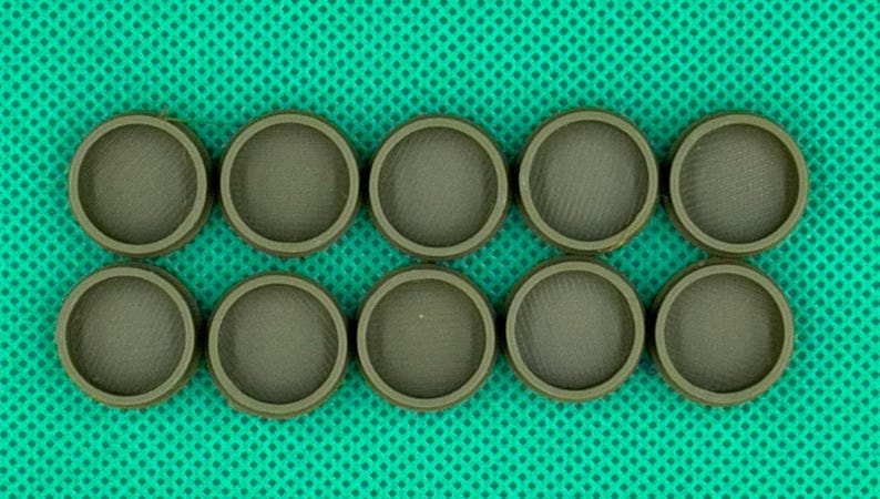May include: Ten circular, olive-green plastic caps arranged in two rows of five. Each cap features a raised outer rim and a recessed, textured center surface with a fine, cross-hatched pattern visible across the flat interior base.