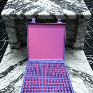 May include: An open, rectangular, pink and purple case with a grid of circular holes. The case is on a marble surface. The case has a purple frame and a pink interior. The case is open, revealing a tray with many circular holes.