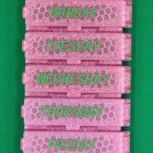 May include: A pink glittery seven-compartment pill organizer with a honeycomb texture. Each rectangular section features a hinged lid labeled with the days of the week in green text, starting with SUNDAY and ending with SATURDAY.