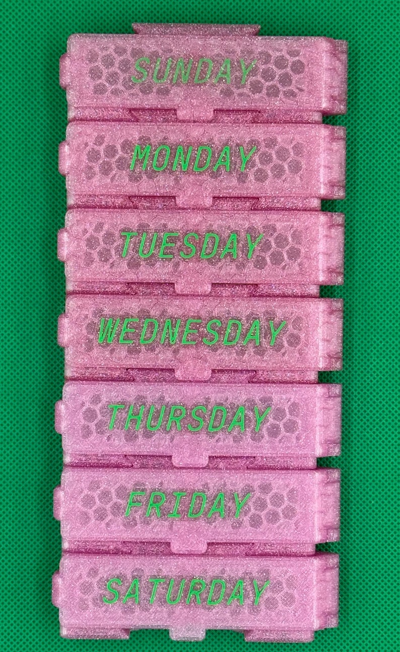 May include: A pink glittery seven-compartment pill organizer with a honeycomb texture. Each rectangular section features a hinged lid labeled with the days of the week in green text, starting with SUNDAY and ending with SATURDAY.