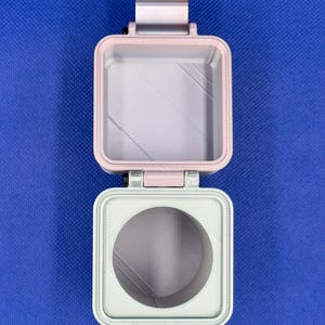 May include: A square, hinged container featuring a light pink top section with a square interior and a light green bottom section with a circular opening. The two parts are connected by a metallic hinge, displaying a layered, textured finish.