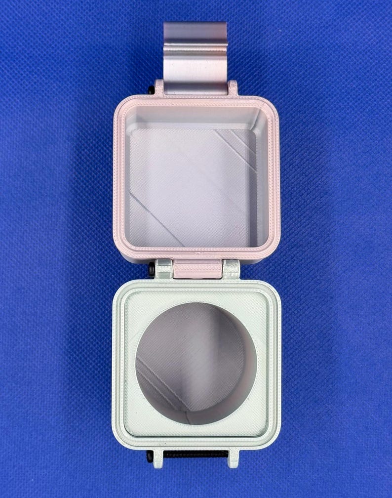 May include: A square, hinged container featuring a light pink top section with a square interior and a light green bottom section with a circular opening. The two parts are connected by a metallic hinge, displaying a layered, textured finish.