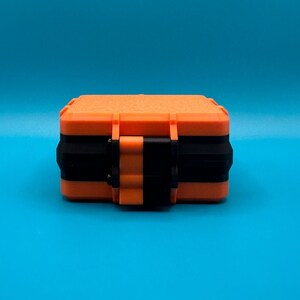 May include: A rectangular storage case featuring a bright orange textured exterior with a contrasting black horizontal band and a central orange and black latch mechanism, designed for secure closure and protection of small items.