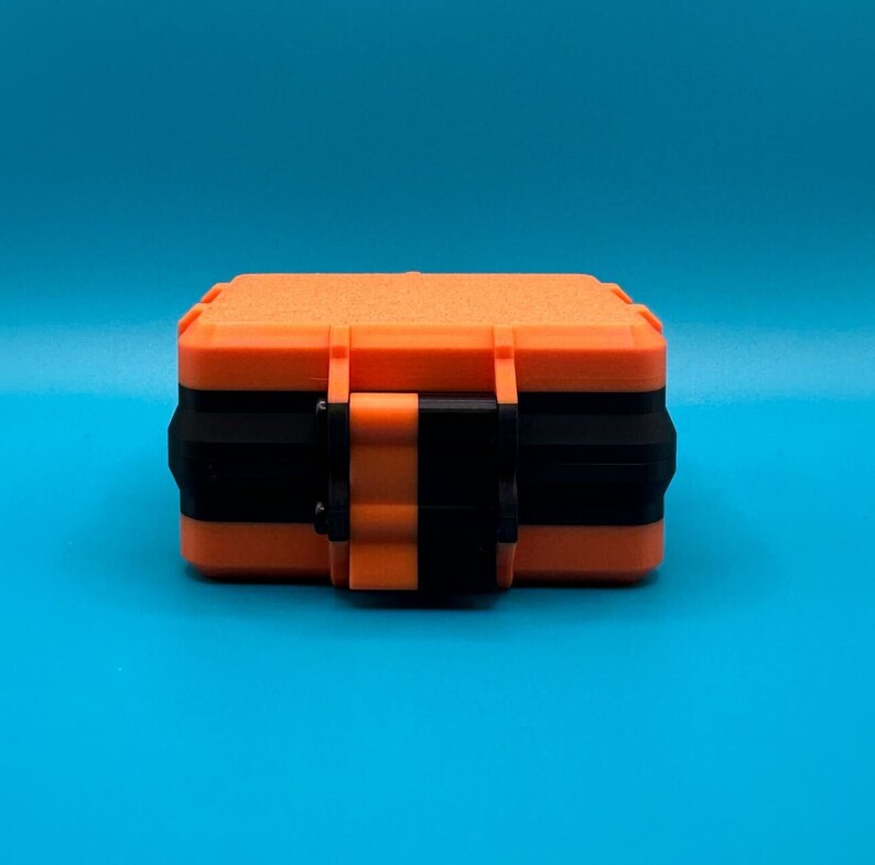 May include: A rectangular storage case featuring a bright orange textured exterior with a contrasting black horizontal band and a central orange and black latch mechanism, designed for secure closure and protection of small items.