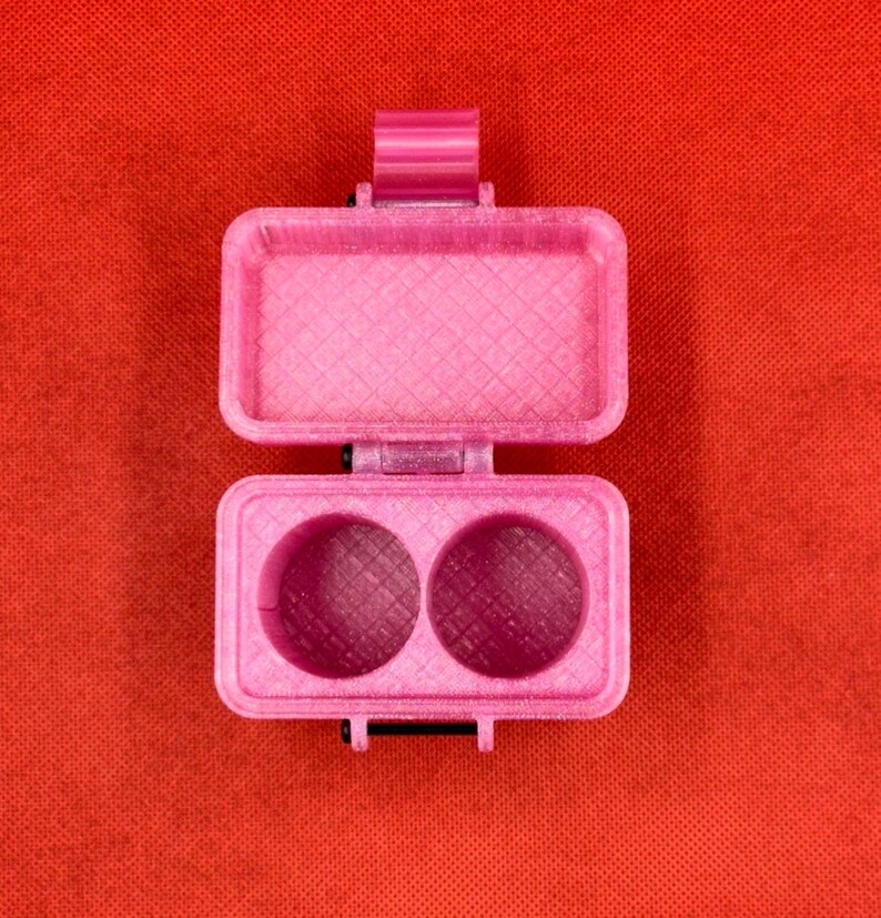 May include: A pink plastic container with two compartments, each with a circular opening. The container has a textured surface and a hinged lid.