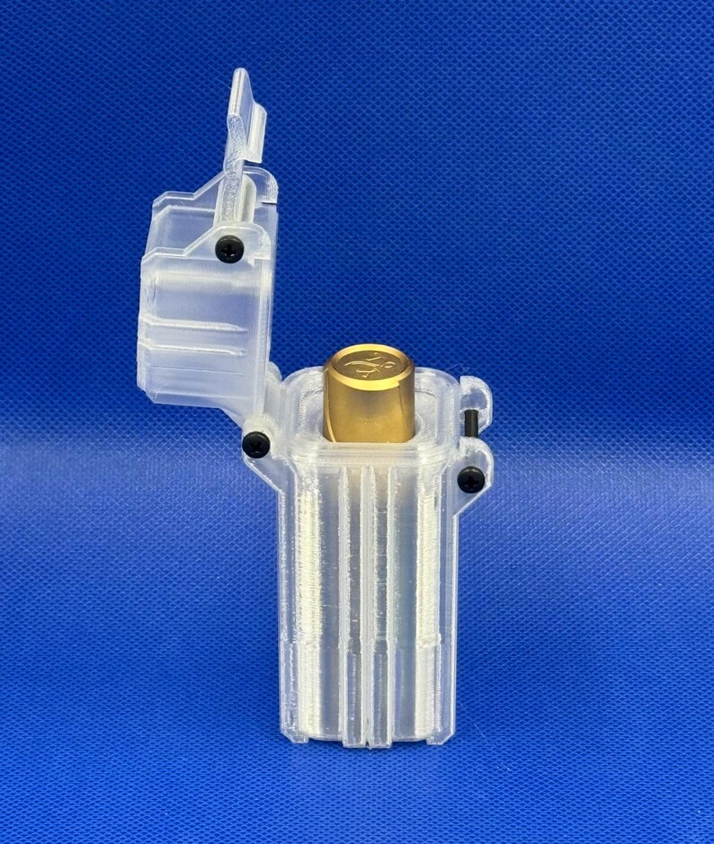 Puede incluir: Un estuche de almacenamiento transparente de pl&aacute;stico impreso en 3D con una tapa abatible sujeta por tornillos negros. Dentro del recipiente rectangular vertical se encuentra un objeto cil&iacute;ndrico de color dorado con un logo.