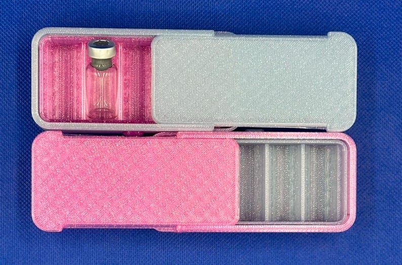 May include: Two rectangular sliding cases with a textured finish. The top case is silver with a bright pink interior containing a small clear glass vial with a metallic cap. The bottom case is bright pink with a silver interior featuring dividers.