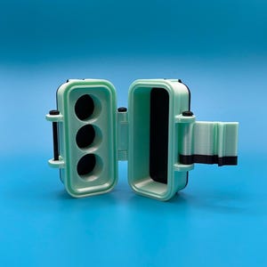Puede incluir: Un estuche de almacenamiento impreso en 3D de color verde menta y negro con tapa abatible. El interior del lado izquierdo presenta tres compartimentos circulares, mientras que el lado derecho tiene una cavidad rectangular profunda.