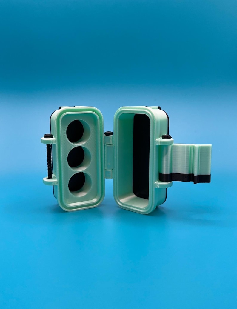Puede incluir: Un estuche de almacenamiento impreso en 3D de color verde menta y negro con tapa abatible. El interior del lado izquierdo presenta tres compartimentos circulares, mientras que el lado derecho tiene una cavidad rectangular profunda.