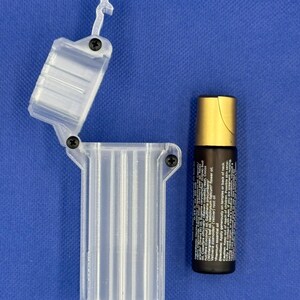 Puede incluir: Un estuche de almacenamiento rectangular y transparente de pl&aacute;stico con bisagra, tres ranuras verticales internas y tornillos negros, junto a un frasco de aceite esencial con aplicador de bola, tapa dorada y etiqueta.