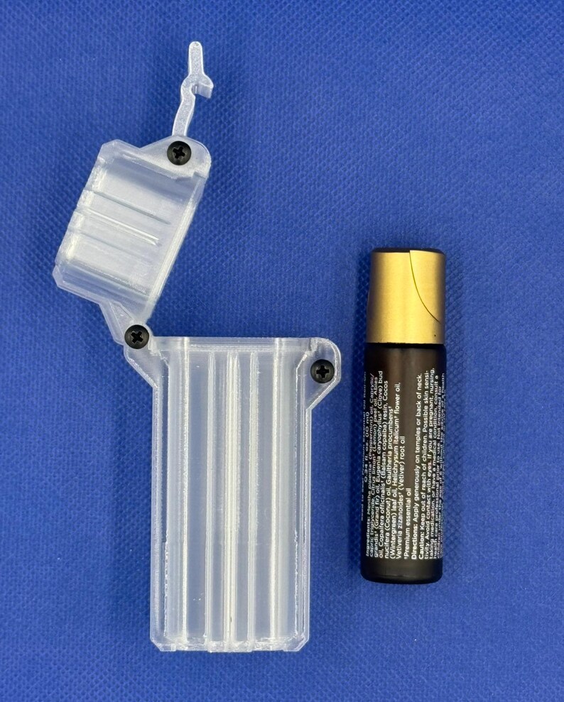 Puede incluir: Un estuche de almacenamiento rectangular y transparente de pl&aacute;stico con bisagra, tres ranuras verticales internas y tornillos negros, junto a un frasco de aceite esencial con aplicador de bola, tapa dorada y etiqueta.