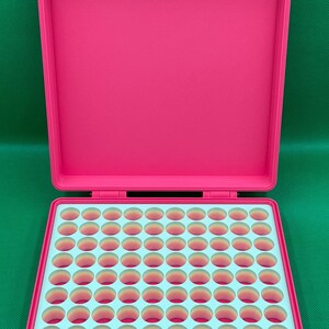 May include: A bright pink plastic storage case with a hinged lid and two latches. The interior features a light blue grid insert with one hundred circular cutouts, each revealing a gradient of pink and orange colors at the base.