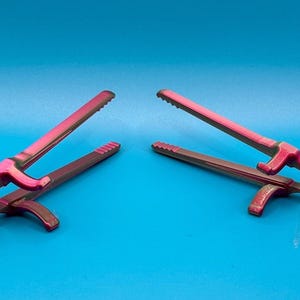 May include: Two kitchen tongs featuring a dual-tone gradient finish that shifts between vibrant magenta and deep metallic green. Each tool has a serrated gripping edge, a central pivot point, and a curved handle design for manual operation.