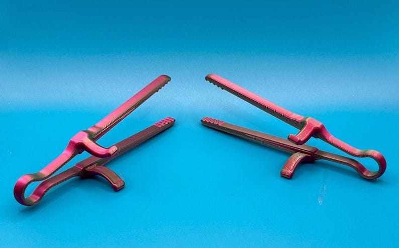 May include: Two kitchen tongs featuring a dual-tone gradient finish that shifts between vibrant magenta and deep metallic green. Each tool has a serrated gripping edge, a central pivot point, and a curved handle design for manual operation.