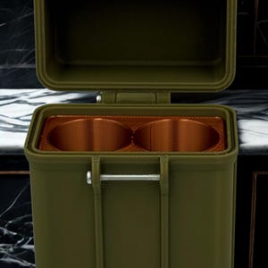 May include: An olive green rectangular storage container with an open hinged lid. The interior features two circular copper-colored compartments. A metallic silver pin secures the front, and a copper-colored strap is attached to the top lid.
