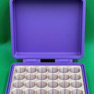 May include: A purple rectangular storage case with two top latches and a hinged lid. The interior features a white insert with a grid of thirty-two circular cylindrical compartments arranged in four rows of eight for organizing small items.