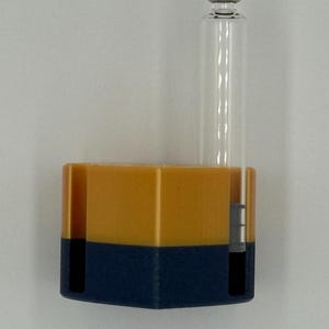 May include: A clear glass vial with a silver cap is attached to a two-tone container. The container is a combination of yellow and navy blue. The vial is cylindrical and the container is rectangular.