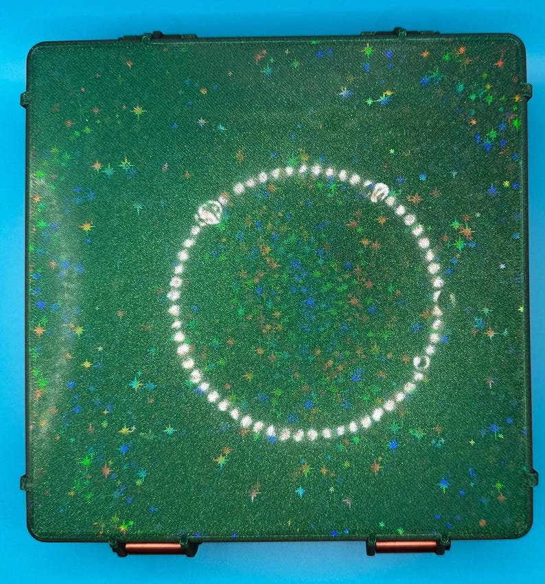 May include: A square, dark green storage case with a textured surface featuring embedded holographic star-shaped glitter. A circular arrangement of small, clear, reflective beads is centred on the lid of the case.