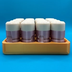 May include: A set of cylindrical containers with cream-colored tops and mauve-colored bases, arranged in a grid pattern within a rectangular orange tray. Each container features a smooth, layered construction with a distinct horizontal seam.