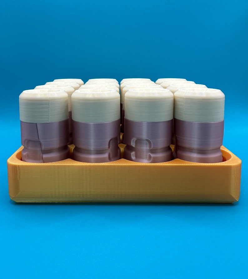 May include: A set of cylindrical containers with cream-colored tops and mauve-colored bases, arranged in a grid pattern within a rectangular orange tray. Each container features a smooth, layered construction with a distinct horizontal seam.