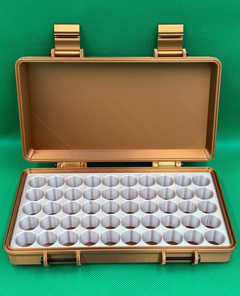 May include: A rectangular, metallic bronze-coloured storage case with an open lid. The interior features a white insert with a grid of fifty-six circular compartments designed for organising small cylindrical items in a structured layout.