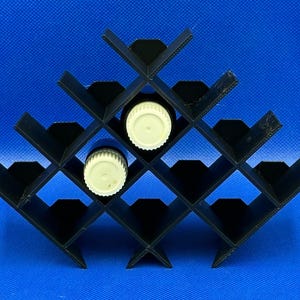 Puede incluir: Estante de plástico negro con 16 ranuras para guardar botellas de aceite esencial. Dos tapas de botellas blancas son visibles en las ranuras.