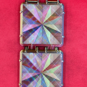 May include: Two square, iridescent components featuring a geometric starburst pattern of fine, etched lines. Each piece has a clear, reflective surface that shifts in color, connected by two brown, striped ribbon tabs at the top edge.