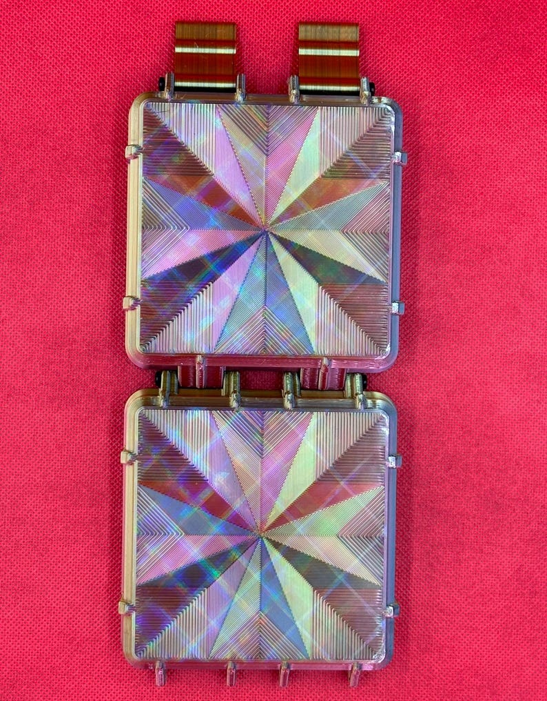 May include: Two square, iridescent components featuring a geometric starburst pattern of fine, etched lines. Each piece has a clear, reflective surface that shifts in color, connected by two brown, striped ribbon tabs at the top edge.