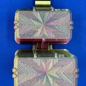 May include: A 3D printed rectangular container with a hinged lid, featuring a metallic gold finish and a multi-colored iridescent star pattern on the top surface. The object has a textured, layered appearance with visible horizontal lines.