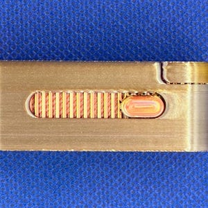 May include: A rectangular, metallic gold-toned object featuring a horizontal recessed channel. Inside the channel is a textured, ribbed copper-coloured sliding mechanism with a small, oval-shaped button positioned on the right side.